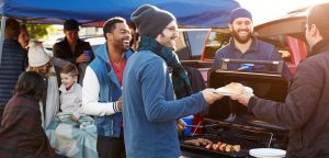 Several people standing and sitting around a Tailgate Party in Action, grilling food and drinking beverages.