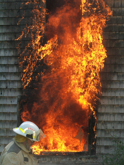 Firefighter standing in front of a house fire with flames leaping out of the window
