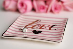 Small pink and white striped square dish with love and a heart drawn on it holding a diamond ring in front of pink roses