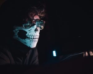 Male wearing sunglasses wearing a skeleton mask over his mouth, appears to be looking a code on a computer screen from the reflection in the sunglasses