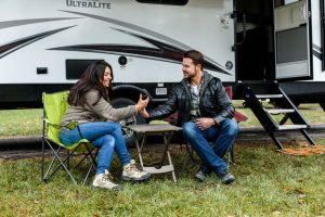 Male and female sitting in camping chairs in front of an RV camper looking at something on a mobile device
