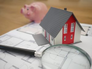 Home Inspection, Small white and red plastic toy house with grey roof on sitting on floor plan drawings next to a magnifying glass with a small toy pink piggy bank nearby.