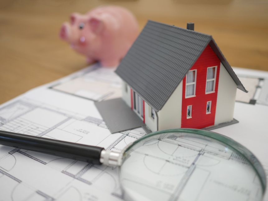 Small white and red plastic toy house with grey roof on sitting on floor plan drawings next to a magnifying glass with a small toy pink piggy bank nearby.