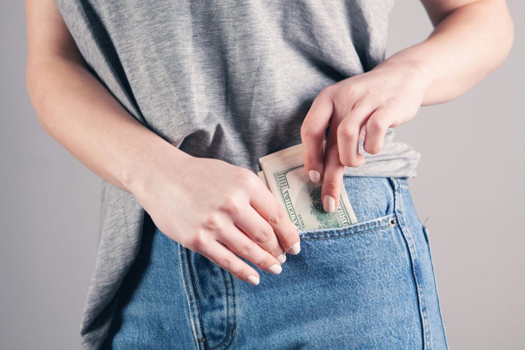Light colored person with gray t-shirt on and blue jeans stuffing hundred dollar bills into front pocket of jeans.